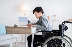 Remote education concept. Focused disabled teenage boy in wheelchair studying online from home, using laptop