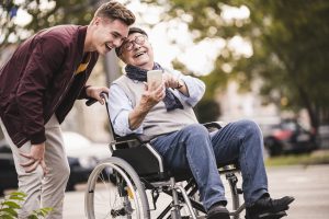 Laughing senior man in wheelchair and his adult grandson looking together at smartphone having fun