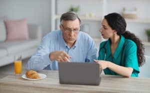 Young nurse showing senior man in wheelchair how to use laptop at retirement home