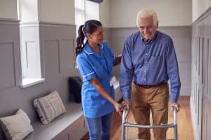 Senior Man At Home Using Walking Frame Being Helped By Female Care Worker In Uniform